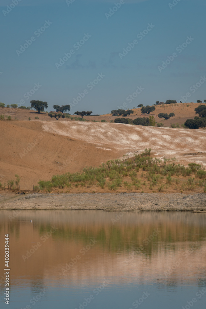 Desert like hill landscape with reflection on the water on a dam lake reservoir with blue sky in Terena, Portugal