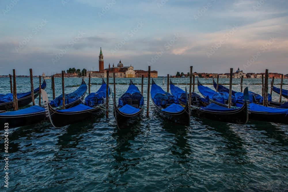 Docked gondola float in the waters near St Mark's Square, with the bell tower of the Church of San Giorgio Maggiore in the background in Venice, Italy at sunset. 