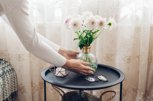 Woman puts vase with flowers dahlias on table. Housewife taking care of coziness in apartment. Interior and decor