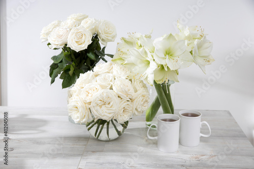 White roses, lily, in round vases with two cups of coffee on the table for a special occasion as a kitchen decoration.
