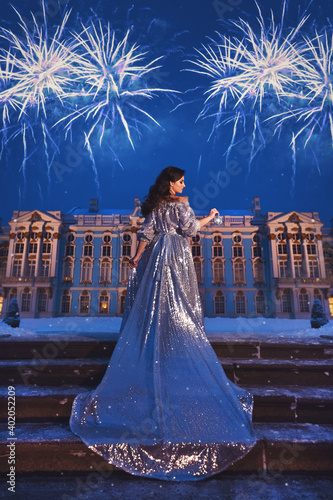 Beautiful girl in a shiny silver dress with a long train standing on the stairs against the background of the palace, the night sky and magnificent fireworks