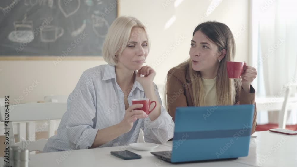 Confident young Caucasian women gossiping in lunchroom on break. Portrait of two beautiful colleagues talking and drinking tea or coffee sitting in lunchroom with laptop. Lunch and rumors. Confident young Caucasian women gossiping in lunchroom on break. Portrait of two beautiful colleagues talking and drinking tea or coffee sitting in lunchroom with laptop. Lunch and rumors.