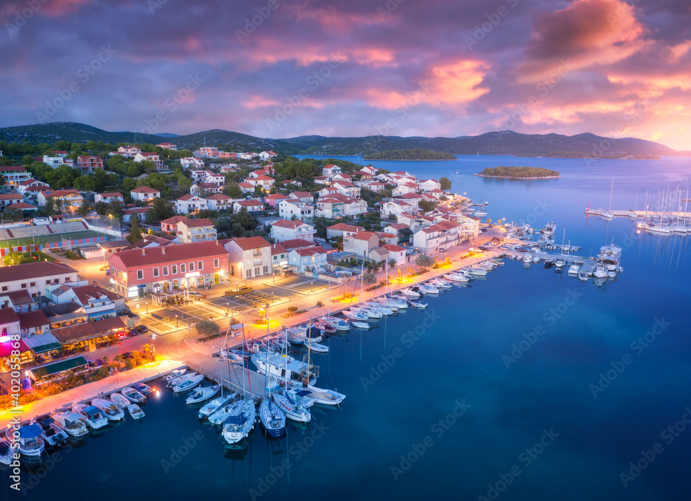 Aerial view of boats and yachts in port and city at night. Summer landscape with city lights, buildings, illuminated streets, mountain, motorboats, blue sea, colorful sky at sunset. Top view. Croatia