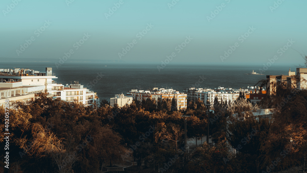 An evening cityscape of a dormitory area near the sea with multiple multi-story residential buildings lit by warm sunset light and a park zone in the foreground, Oeiras district of Lisbon, Portugal