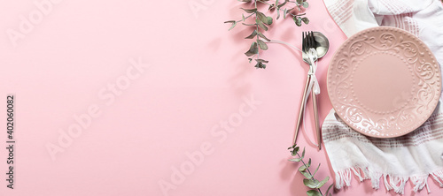 Long wide banner with pink empty plate, white napkin, cutlery set with bow and decorative eucalyptus twigs on pink pastel background. Festive table setting for Valentine’s Day or Easter concept.
