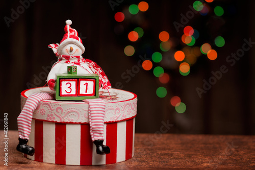 486.	A snowman figure is sitting on a decoupage box and holding a smaller box with two cubes forming a number thirty-one. A beautiful bokeh made with Christmas lights is in the background. photo