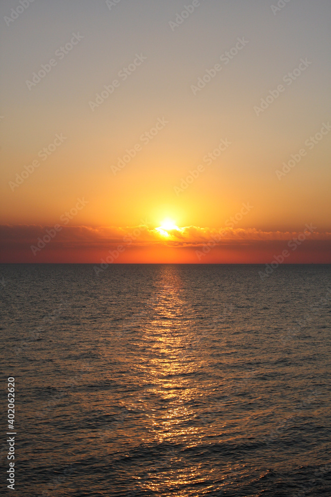 Sunset. Sun sets behind sea horizon, turning clouds red and orange. Vertical orientation