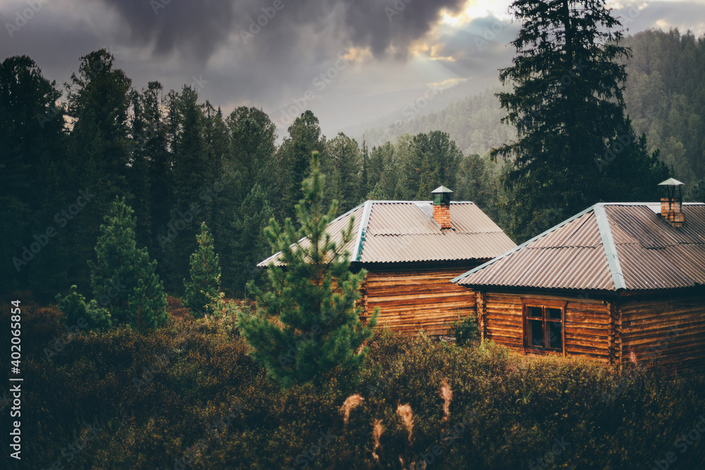 A beautiful rainy rural scenery in the mountains with shallow depth of field and selective focus on two small wooden cabins in deep taiga forest with particles of rain everywhere, dramatic stormy sky