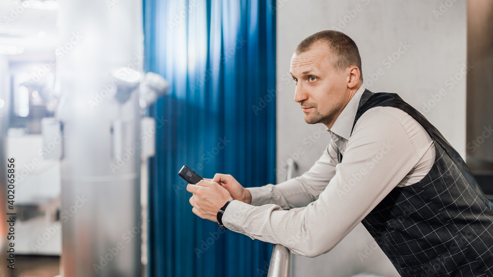 Fototapeta premium A man entrepreneur in a business suit and with a cellphone in hands leaning against the balcony railing in a modern office coworking area; a businessman in a formal costume using a cellphone indoors