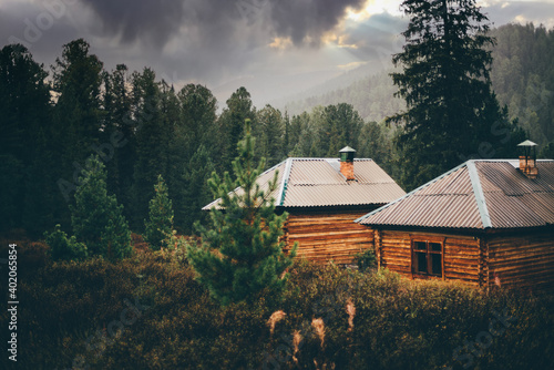 A beautiful rainy rural scenery in the mountains with shallow depth of field and selective focus on two small wooden cabins in deep taiga forest with particles of rain everywhere, dramatic stormy sky