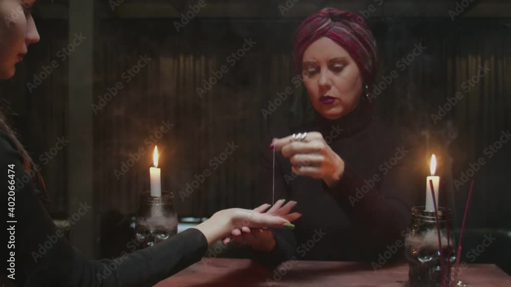 Senior woman fortune teller uses needle with thread to tell the future to her young client Senior woman fortune teller uses needle with thread to tell the future to her young client