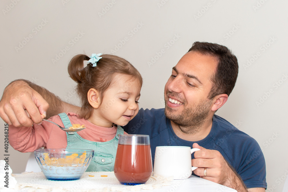 Handsome young father together with cute baby daughter having breakfast together. Dad and child eating corn flakes morning at home during quarantine of pandemic Coronavirus. Happy fathers day concept. Handsome young father together with cute baby daughter having breakfast together. Dad and child eating corn flakes morning at home during quarantine of pandemic Coronavirus. Happy fathers day concept.