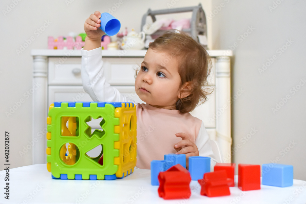 Cute adorable baby girl solves sorter puzzle. Toddler Having Fun with educational toys. Kid playing in nursery with nordic stylish. White scandinavian interior for children's room. Cute adorable baby girl solves sorter puzzle. Toddler Having Fun with educational toys. Kid playing in nursery with nordic stylish. White scandinavian interior for children's room.