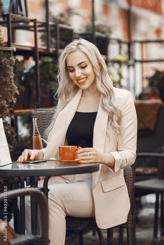Lady drinks a coffee. Woman sitting at the table. Blonde in a white suit.