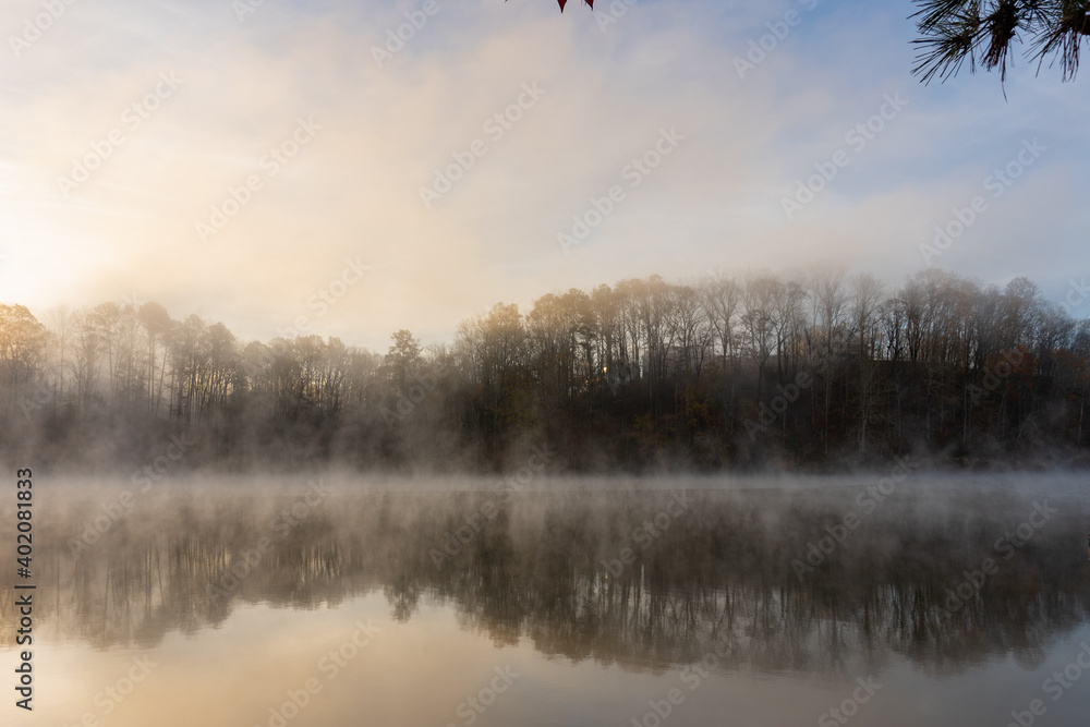 Fototapeta premium Fog rises on the water at sunrise on Lake Lanier in Georgia with a reflection of trees in the water in winter