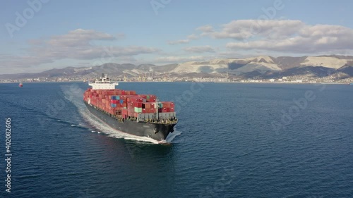 Aerial view of large cargo ship with red containers floating on sea waves on warm sunny day. Concept of work on sea delivery of goods and cargo