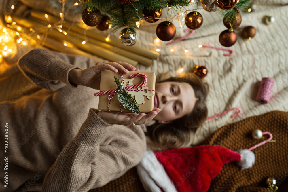 Fototapeta premium Christmas composition. Woman wrapping gift box in craft paper near fir tree, next lying santa hat, candy canes. new year concept. Xmas celebration 2021. Flat lay, top view, overhead