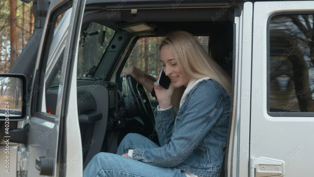 Young blonde woman sits on the passenger seat in the car jeep and talks on phone in autumn forest Young blonde woman sits on the passenger seat in the car jeep and talks on phone in autumn forest