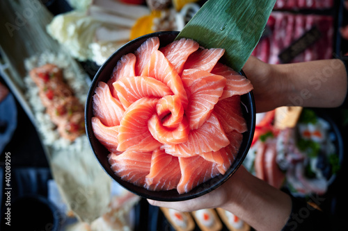 Hands holding a bowl of salmon sashimi on top of a table full of japanese food. Top view.
