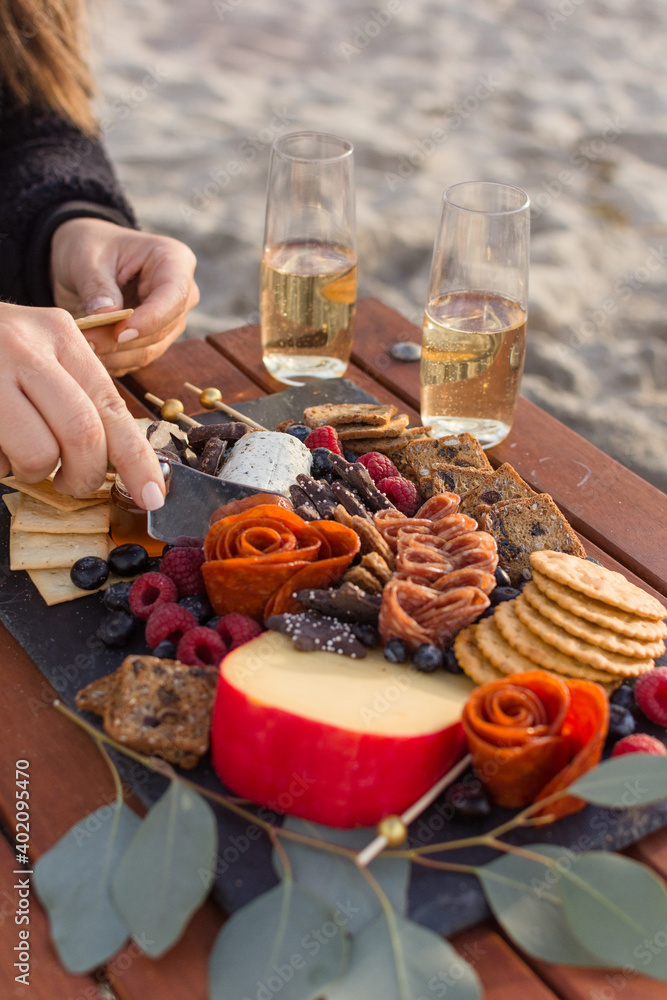 Woman's hand cutting into soft goat cheese onto a cracker from a grazing board of charcuterie on a wood table with two glasses of champagne with blurry beach sand in the background.