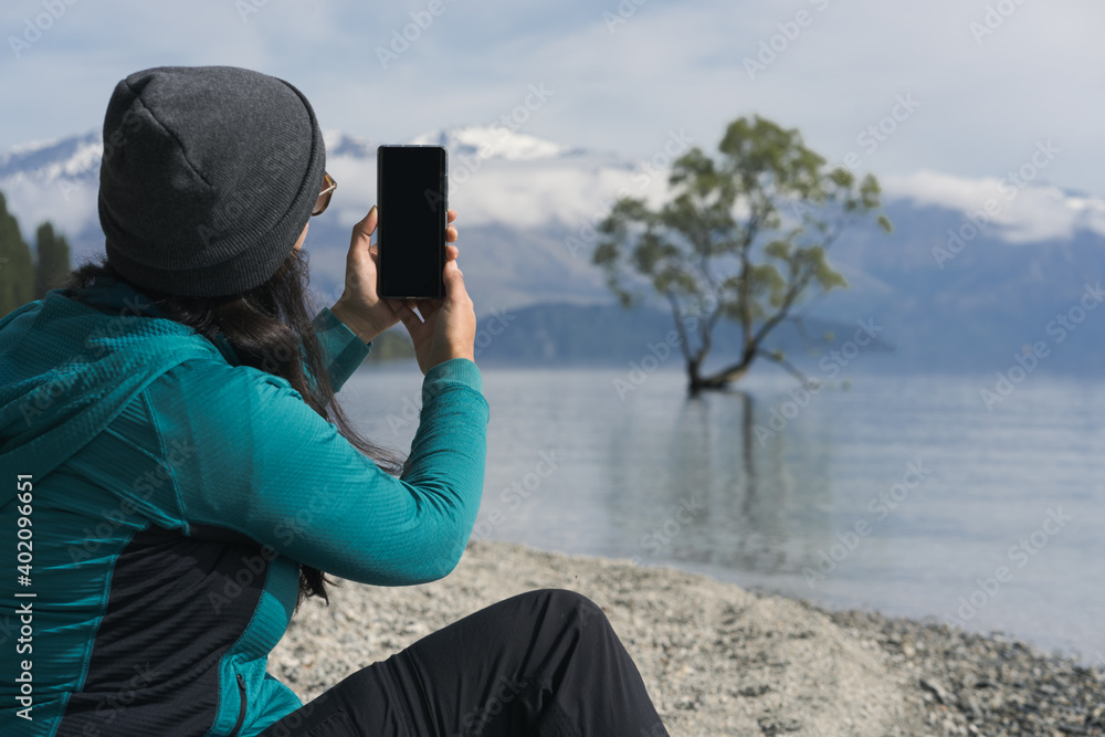 Obraz premium Woman taking picture with smartphone at iconic Wanaka Tree. New Zealand