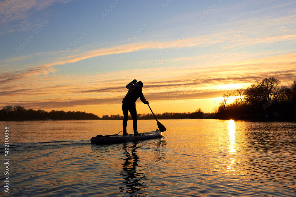 Silhouette of woman paddle on stand up paddle boarding (SUP) on quiet winter river at sunset