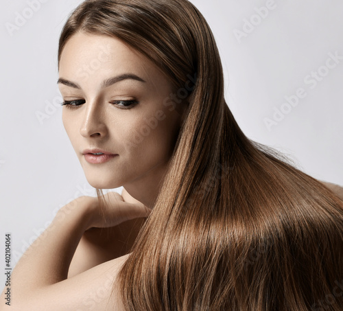 Profile of young woman lying on floor on elbows and looking at her long silky straight hair falling over her shoulder after haircare spa treatment over grey background. Haircare, beauty, wellness