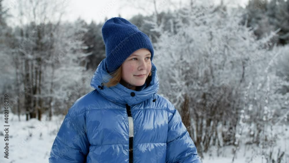 Cute smiling girl walking in snowy winter park. Adorable happy young girl looking aside while walking between snow-covered trees at wintertime Cute smiling girl walking in snowy winter park. Adorable happy young girl looking aside while walking between snow-covered trees at wintertime