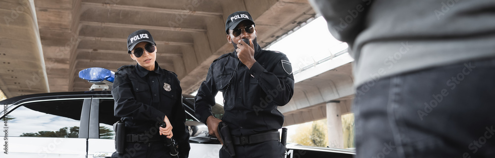 african american police officer talking on radio set near colleague taking out police bat with blurred offender on foreground, banner. african american police officer talking on radio set near colleague taking out police bat with blurred offender on foreground, banner.