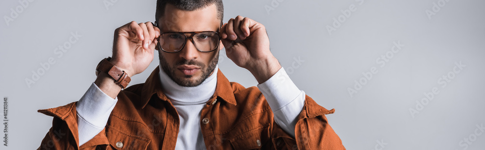Serious stylish man adjusting eyeglasses and looking at camera isolated on grey, banner - obrazy, fototapety, plakaty 