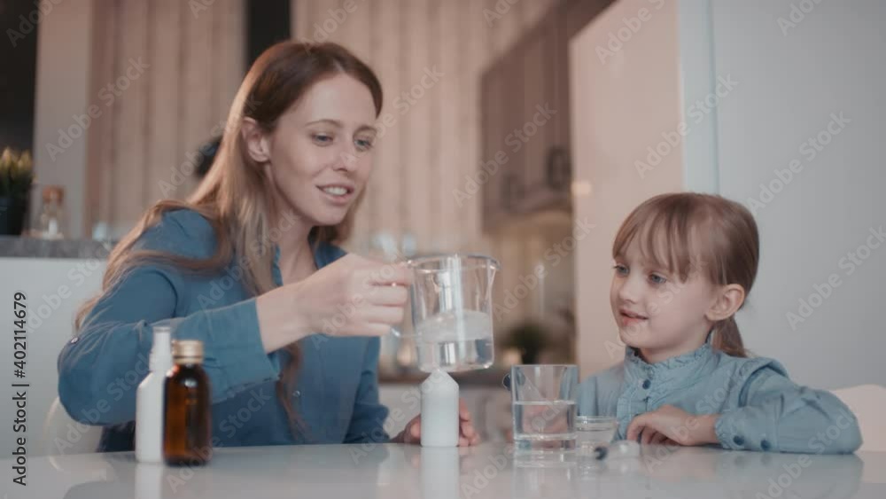 Medium slow-motion shot of enthusiastic mother and little girl staying at home at lockdown making antiseptic using measuring cup with mixed ethanol, hydrogen peroxide and glycerin