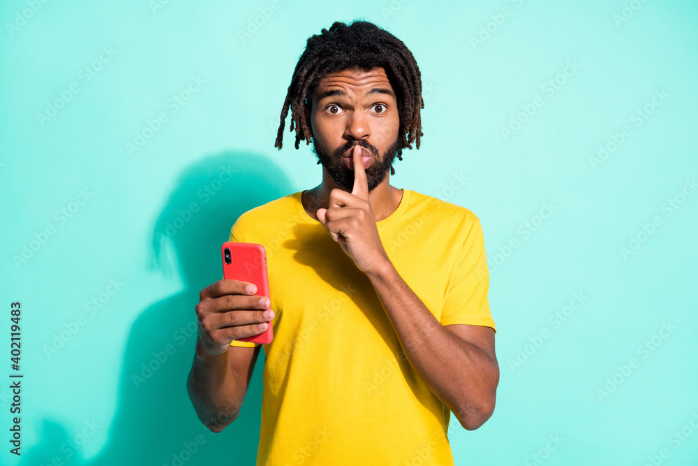 Photo of young attractive african man wear yellow t-shirt hold finger on lips use smartphone isolated on teal color background Photo of young attractive african man wear yellow t-shirt hold finger on lips use smartphone isolated on teal color background