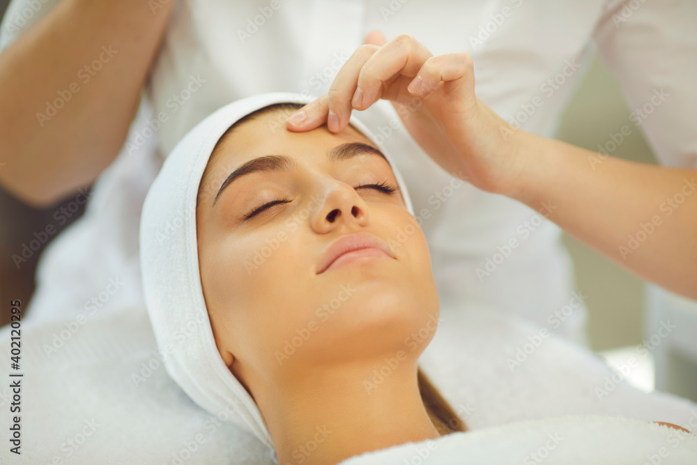 Hands of cosmetologist checking skin elasticity or making facial cleaning for young relaxing woman