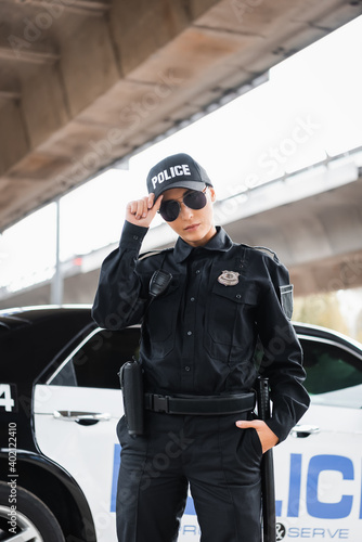 young policewoman with hand in pocket looking at camera near patrol car on blurred background outdoors.