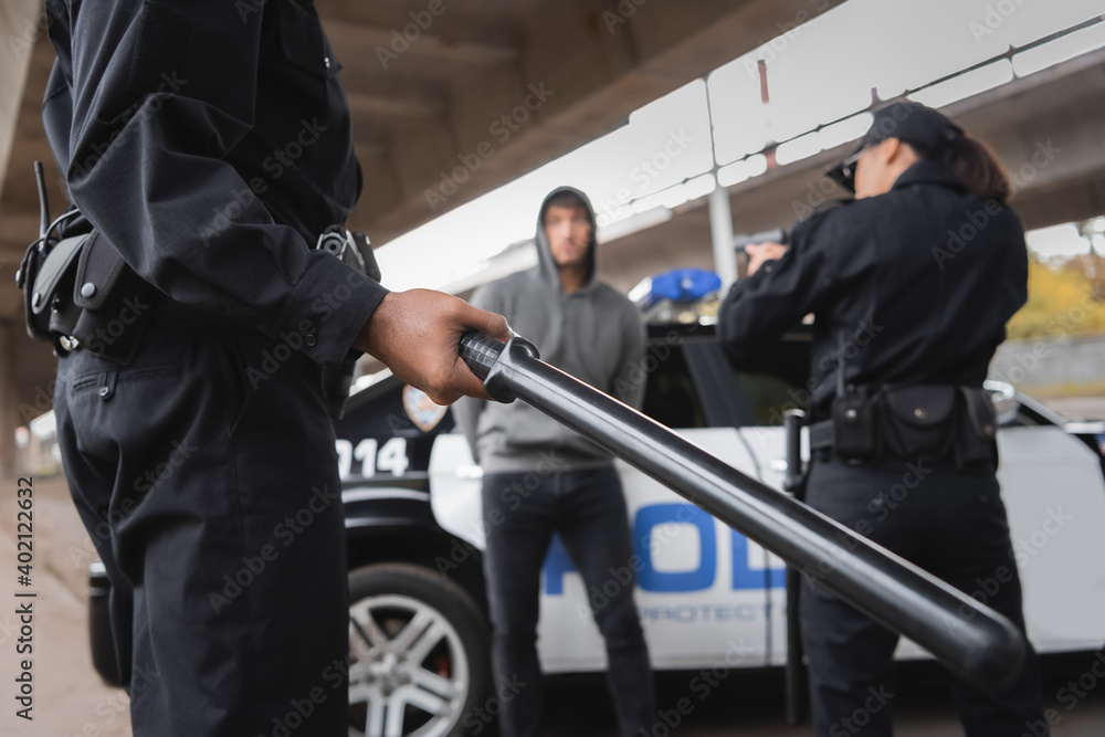 Obraz premium african american police officer holding truncheon near colleague aiming with pistol and offender on blurred background outdoors.