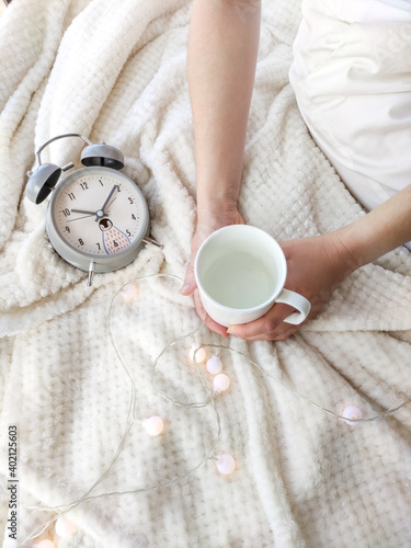 Two hands holds a white cup and alarm clock on a white plaid in garland lights top view