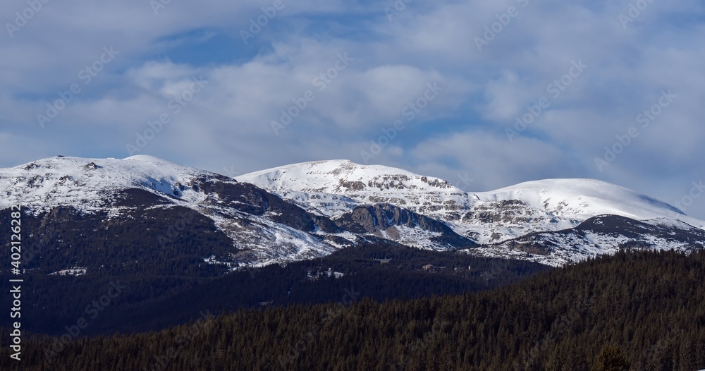 Beautiful winter panoramic natural mountain landscape. Attractive snowy peaks of Rila Mountains, Bulgaria. White clouds in dynamic blue sky, perfect conditions for tourism recreation and winter sports