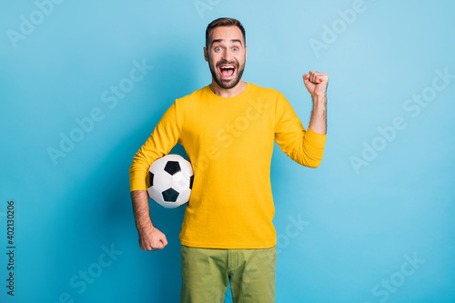 Photo of young happy excited crazy smiling cheerful man with football ball raise fist in victory isolated on blue color background © deagreez