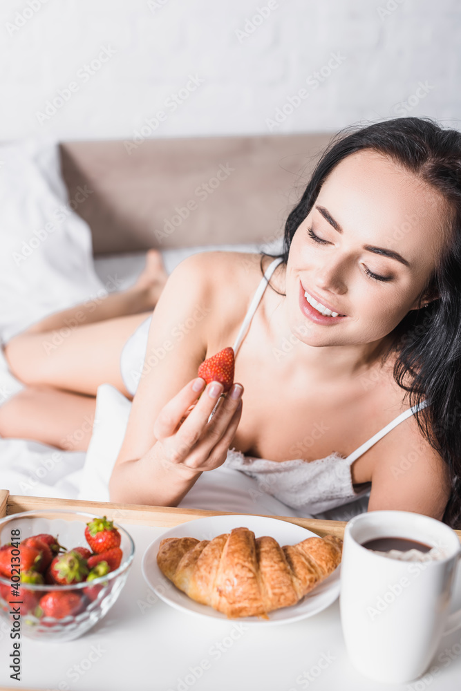  brunette woman having croissant, strawberry and cocoa for breakfast in bed