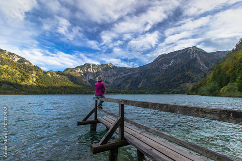 Young girl having a rest by mountain lake, Austria. Female traveler enjoying view of Alps. Wanderlust freedom travel concept. Summer vacation adventure scene. Sitting relaxing woman on her holiday