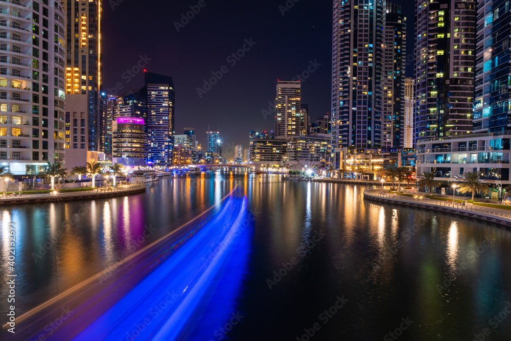 Night view to Dubai Marina panorama, reveals Pier 7, skyscrapers, speed boat light trails and beautiful bridge. Amazing colors reflect on the water.