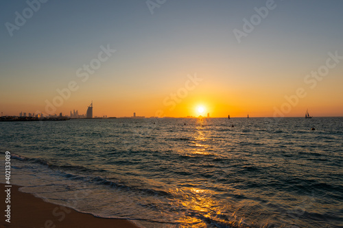 Amazing orange sunset at the beach in Dubai. Beautiful orange colors coming from sun and reflect over Gulf sea waves and shore of UAE. Silhouette of the iconic hotels visible at the horizon line. 