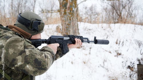 Submachine gun with a silencer during shooting, a shooter with headphones removed in a military color cap on a winter background