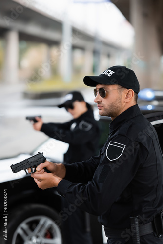 Police officer in sunglasses holding firearm near colleague and car on blurred background outdoors.