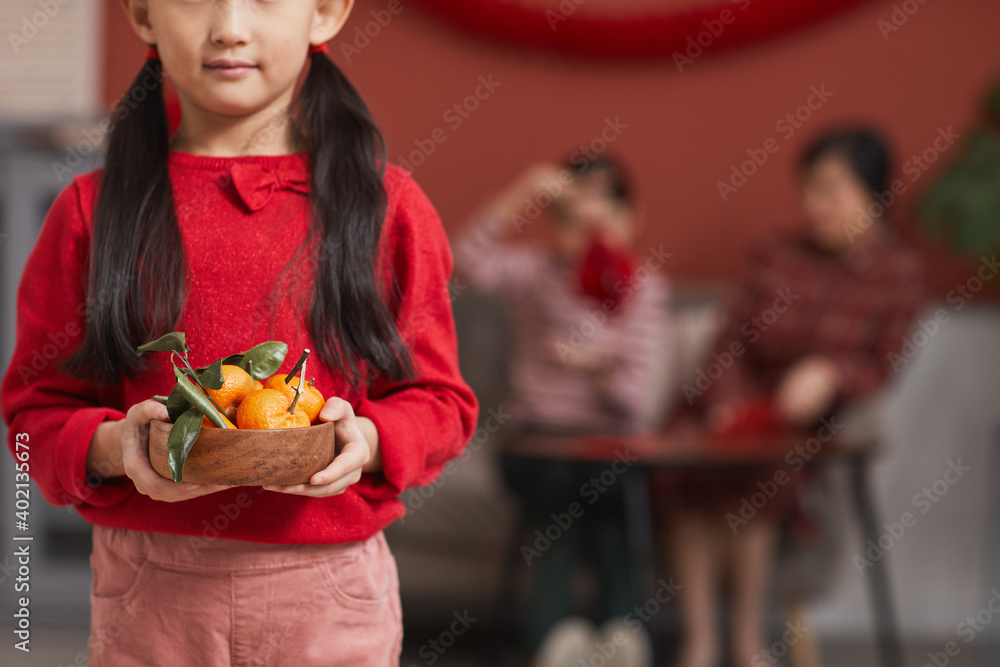 Medium section shot of modern Chinese girl holding wooden bowl full of fresh tangerines, Lunar New Year traditions concept