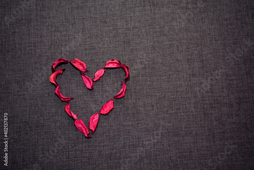 Символ сердца из сухих лепестков красной розы на сером текстильном фоне.