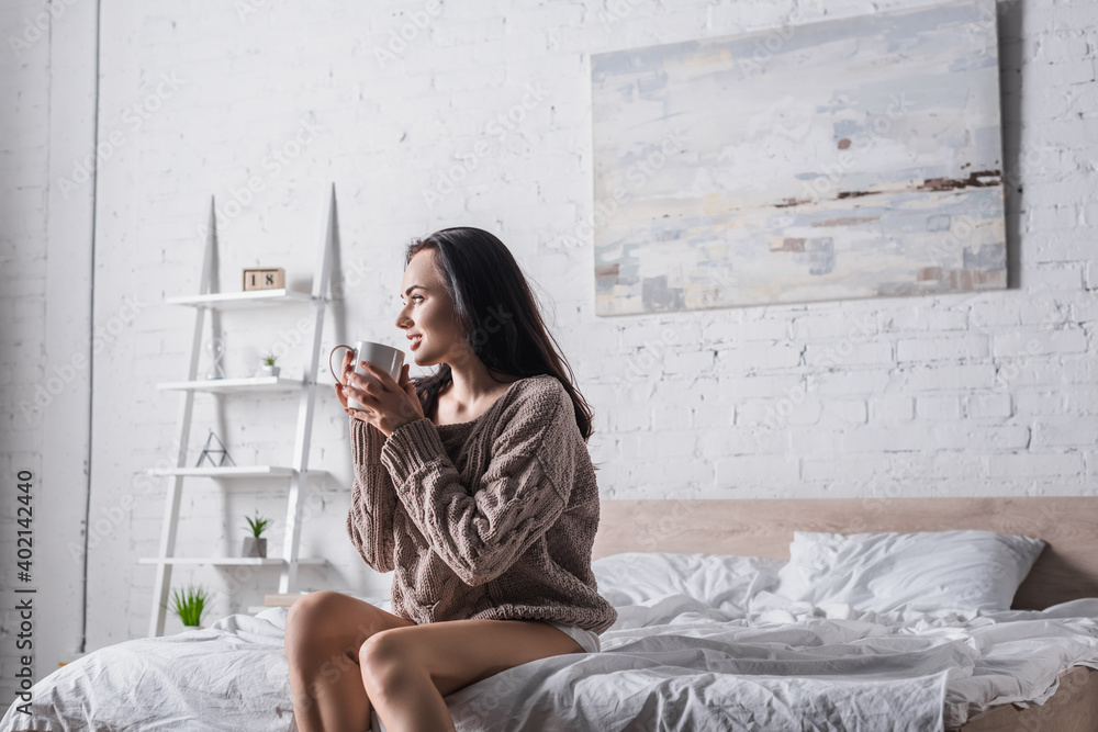 brunette woman in sweater sitting in bed with mug of hot cocoa at morning brunette woman in sweater sitting in bed with mug of hot cocoa at morning