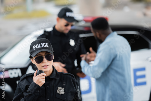 young policewoman talking on radio set with blurred colleague and african american man on background outdoors.