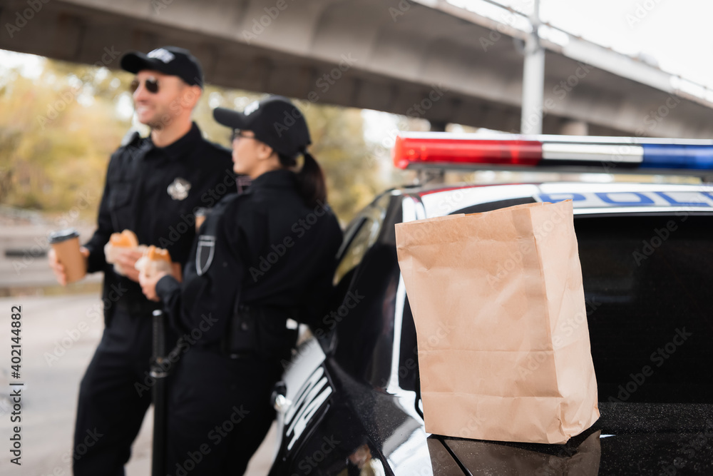 Paper bag on trunk of car near police officers with burgers and coffee to go on blurred background on urban street. Paper bag on trunk of car near police officers with burgers and coffee to go on blurred background on urban street.