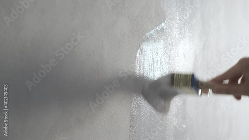 Woman house painter is painting white wall wallpaper in gray using paintbrush, hand closeup. Making renovation in flat, room, house, apartment. Finishing, decoration and construction works.